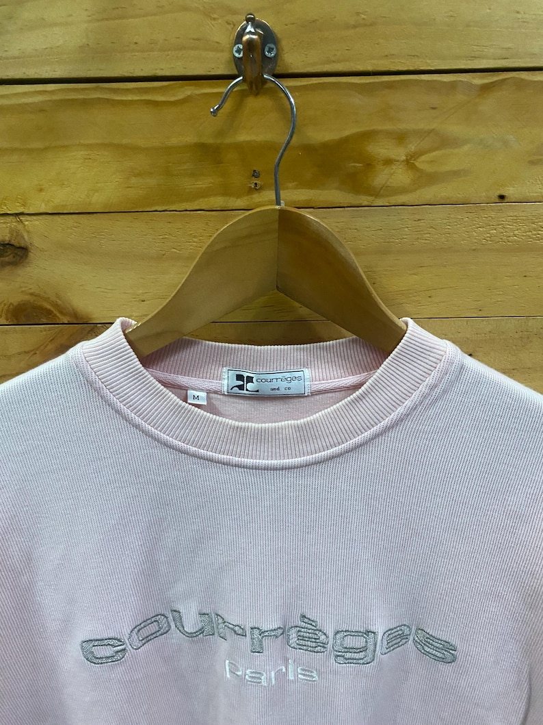 May include: A light pink Courr&egrave;ges Paris sweatshirt with a crew neck. The brand name is embroidered in silver thread across the chest. The sweatshirt is hanging on a wooden hanger against a wood-paneled background.