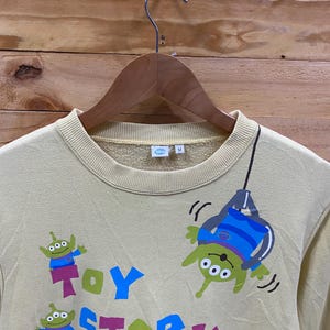 K&ouml;nnte beinhalten: Ein hellgelber Pullover mit einem Toy Story-Motiv. Der Pullover zeigt eine gr&uuml;ne Alienfigur, die an einem Seil h&auml;ngt, mit den Worten "Toy Story" in bunten Buchstaben darunter. Auf der Vorderseite des Pullovers befinden sich auch zwei kleinere Alienfiguren.