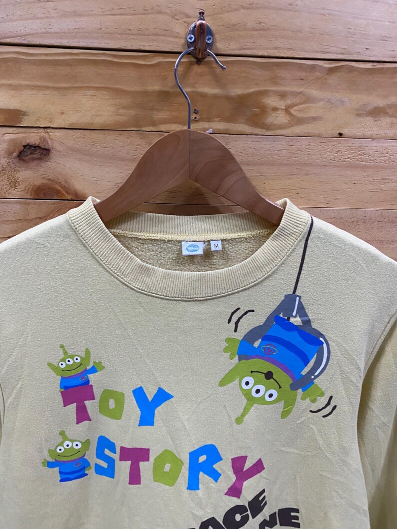 K&ouml;nnte beinhalten: Ein hellgelber Pullover mit einem Toy Story-Motiv. Der Pullover zeigt eine gr&uuml;ne Alienfigur, die an einem Seil h&auml;ngt, mit den Worten "Toy Story" in bunten Buchstaben darunter. Auf der Vorderseite des Pullovers befinden sich auch zwei kleinere Alienfiguren.