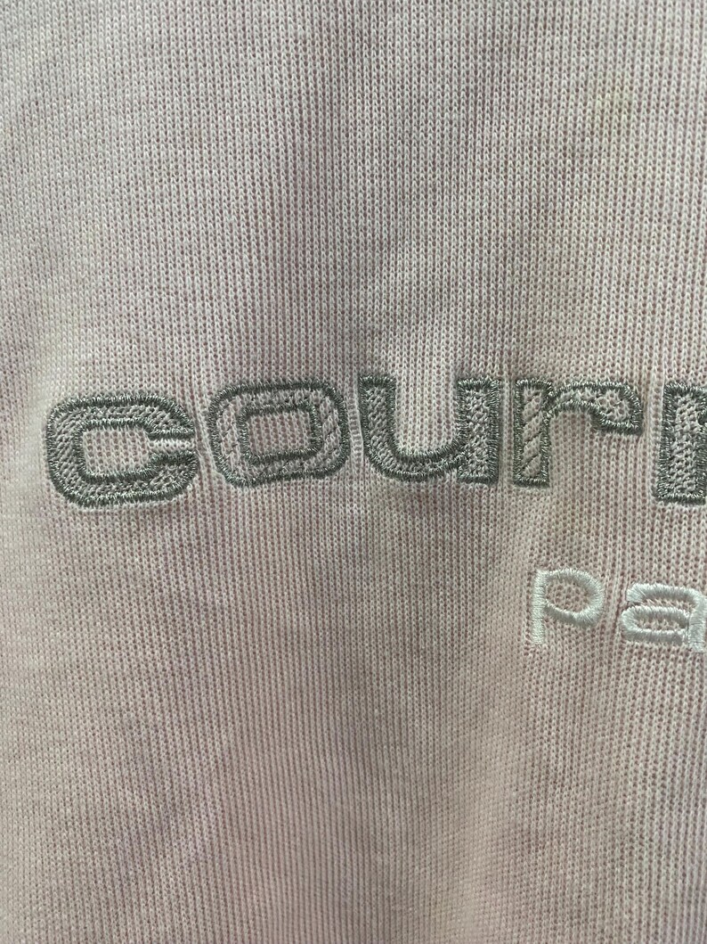May include: Close-up of a light pink fabric with embroidered text. The word "courre" is stitched in a muted green, with "pa" in white below. The fabric has a ribbed texture.