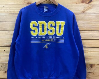 May include: A royal blue crewneck sweatshirt featuring the yellow text SDSU, the words SOUTH DAKOTA STATE UNIVERSITY, and the word JACKRABBITS above a small yellow and blue rabbit mascot graphic. The garment has long sleeves and ribbed cuffs.