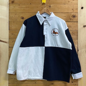 May include: A white and navy blue long-sleeve rugby shirt with a patch on the chest that says "Mainland Nash House Marinas Abu Dhabi".