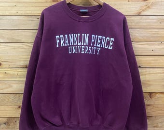 May include: A maroon crewneck sweatshirt with the words "FRANKLIN PIERCE UNIVERSITY" printed in white across the chest. The sweatshirt is hanging on a wooden hanger against a wooden plank background.