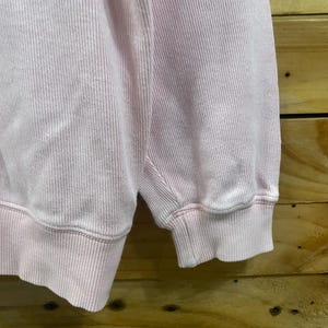 May include: Close-up of a light pink sweatshirt. The fabric has a ribbed texture, with vertical lines running down the material. The cuff of the sleeve is visible, showing the ribbed texture and the hem.