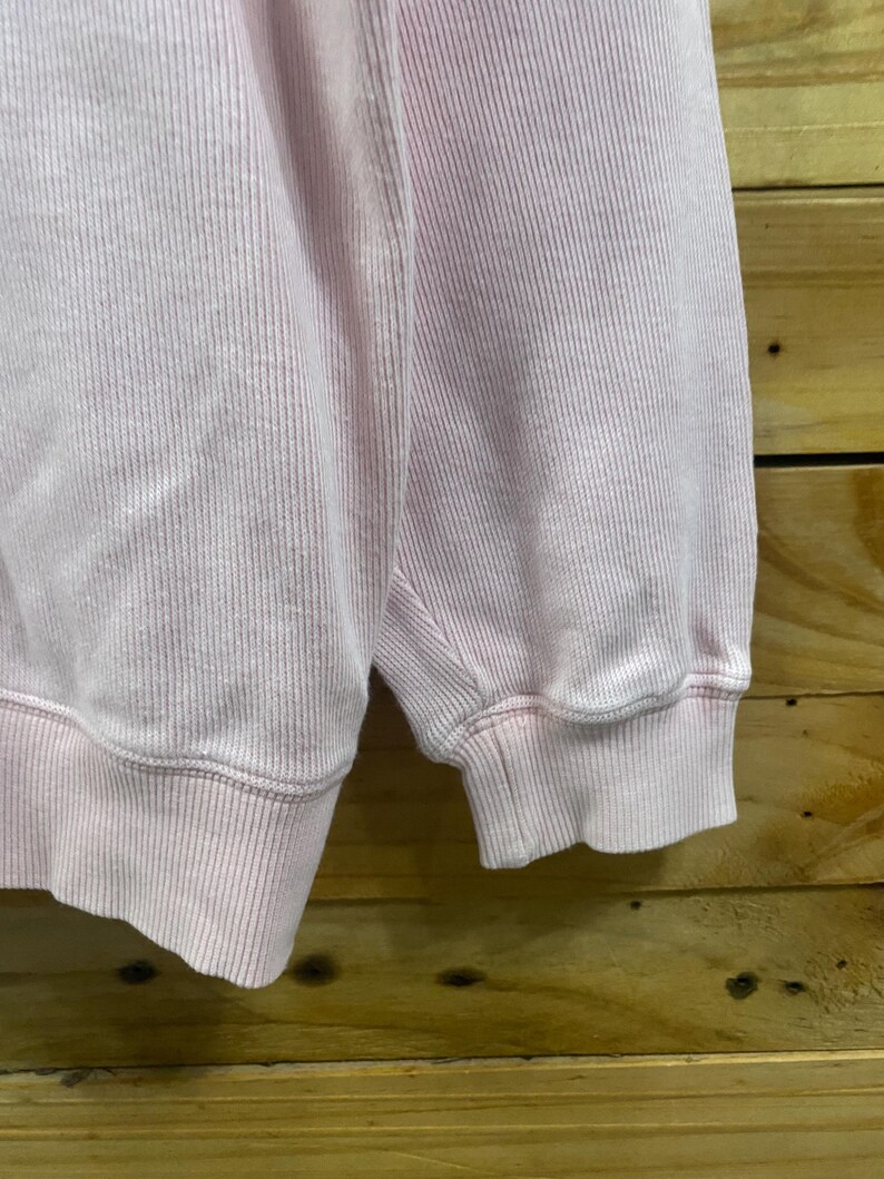 May include: Close-up of a light pink sweatshirt. The fabric has a ribbed texture, with vertical lines running down the material. The cuff of the sleeve is visible, showing the ribbed texture and the hem.