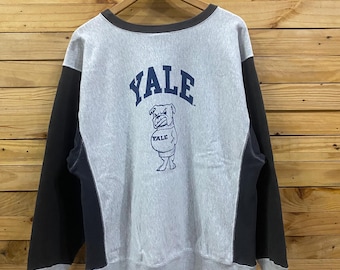 May include: A grey crewneck sweatshirt with dark charcoal side panels and sleeves. The front features the word YALE in navy blue lettering above a graphic of a bulldog wearing a sweater with the word YALE printed on it, all in navy blue ink.