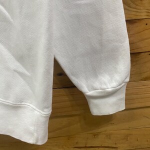 K&ouml;nnte beinhalten: Nahaufnahme eines wei&szlig;en Sweatshirt-&Auml;rmels. Der &Auml;rmel hat einen gerippten Bund und einen Rundhalsausschnitt. Der Stoff scheint ein weiches, strukturiertes Material zu sein. Der Hintergrund ist eine Holzoberfl&auml;che.