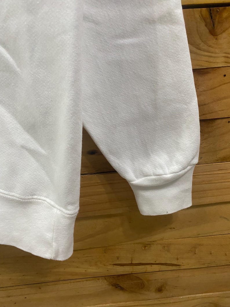 K&ouml;nnte beinhalten: Nahaufnahme eines wei&szlig;en Sweatshirt-&Auml;rmels. Der &Auml;rmel hat einen gerippten Bund und einen Rundhalsausschnitt. Der Stoff scheint ein weiches, strukturiertes Material zu sein. Der Hintergrund ist eine Holzoberfl&auml;che.