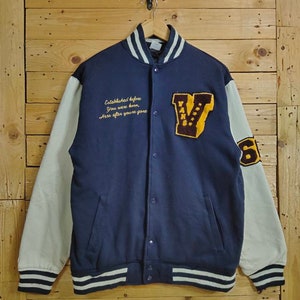 May include: A navy blue varsity jacket with white sleeves and a yellow and brown "V" patch on the chest. The jacket has a white and black striped collar and cuffs. The text "Established before you were born, here after you're gone" is embroidered on the front of the jacket.