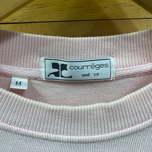May include: Close-up of a light pink Courr&egrave;ges sweatshirt. The label features the brand name and "and co" in a rectangular frame. The size "M" is visible on the left side. The fabric has a ribbed texture around the collar and cuffs.