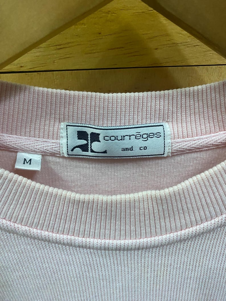 May include: Close-up of a light pink Courr&egrave;ges sweatshirt. The label features the brand name and "and co" in a rectangular frame. The size "M" is visible on the left side. The fabric has a ribbed texture around the collar and cuffs.