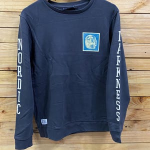 May include: Dark gray sweatshirt with the words "NORDIC" and "DARKNESS" printed vertically in white on the sleeves. A square patch on the chest features a skull design. The sweatshirt is hanging on a wooden hanger.