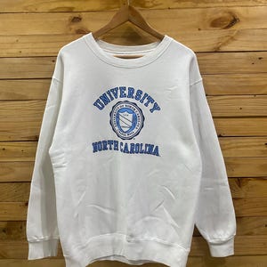 K&ouml;nnte beinhalten: Wei&szlig;es Sweatshirt mit Rundhalsausschnitt und dem Logo der University of North Carolina in Blau. Das Logo ist kreisf&ouml;rmig mit Text und einem Schilddesign. Das Sweatshirt h&auml;ngt an einem Holzb&uuml;gel vor einem Holzgrund.