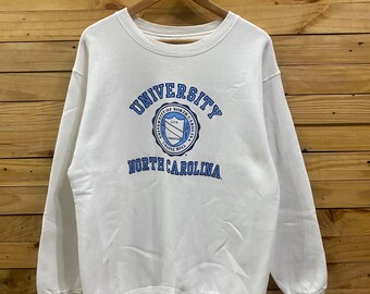 May include: White crew neck sweatshirt featuring the University of North Carolina logo in blue. The logo is circular with text and a shield design. The sweatshirt is hanging on a wooden hanger against a wooden background.
