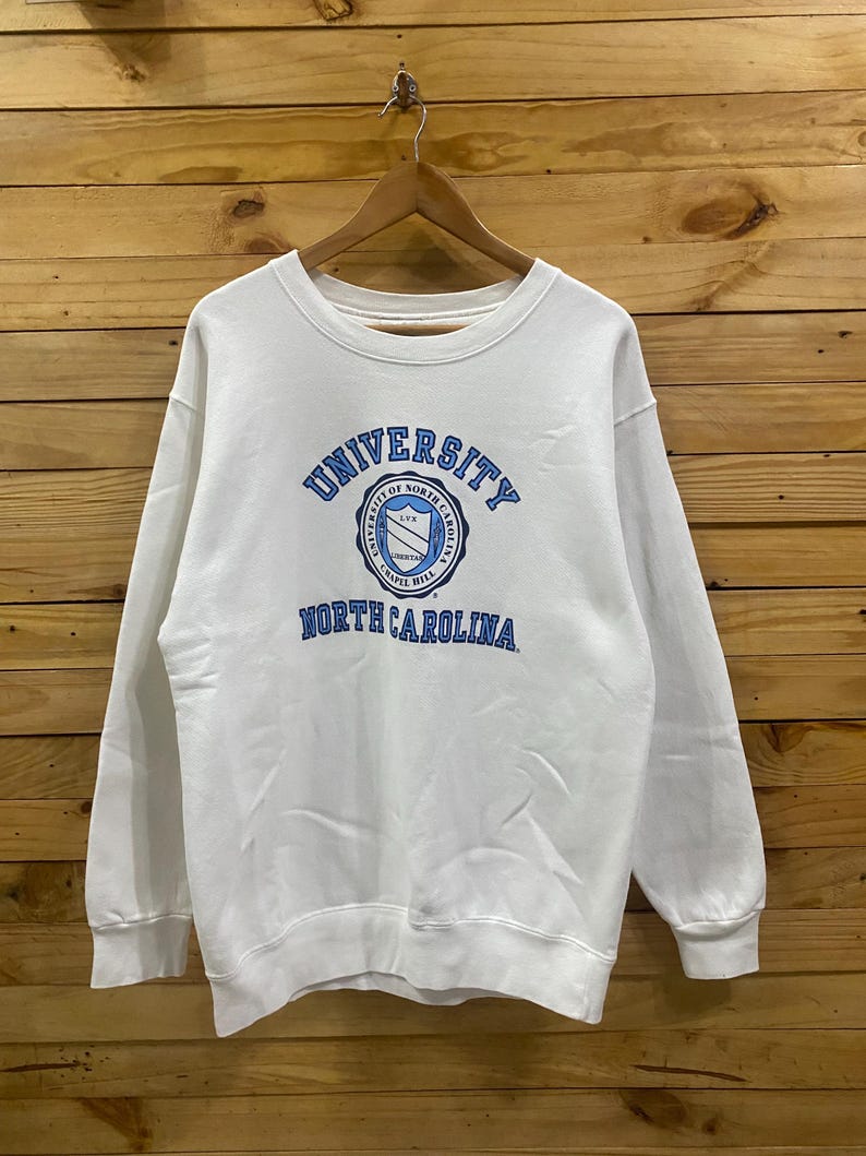 K&ouml;nnte beinhalten: Wei&szlig;es Sweatshirt mit Rundhalsausschnitt und dem Logo der University of North Carolina in Blau. Das Logo ist kreisf&ouml;rmig mit Text und einem Schilddesign. Das Sweatshirt h&auml;ngt an einem Holzb&uuml;gel vor einem Holzgrund.