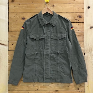 Puede incluir: Una chaqueta de trabajo verde con cierre de botones y dos bolsillos de solapa. La chaqueta tiene un parche de la bandera alemana en cada manga.