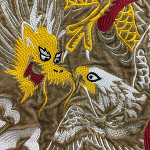 Pu&ograve; includere: Disegno ricamato con un drago giallo con accenti rossi e un'aquila bianca con ali grigie su uno sfondo di tessuto marrone.