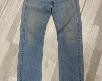 May include: A pair of blue denim jeans featuring a classic five-pocket design, a button fly closure, and copper-toned rivets. The fabric displays a faded wash with light whiskering at the hips and subtle wear along the edges of the pockets and hems.