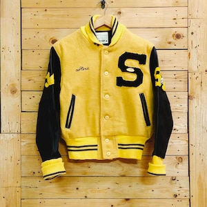 Puede incluir: Una chaqueta de béisbol amarilla y negra con mangas de cuero. La chaqueta tiene una gran "S" en la parte delantera y la palabra "College" bordada en el lado izquierdo.