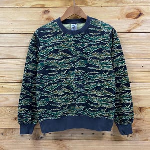 May include: A dark blue sweatshirt with a green, black, and yellow tiger camo print. The sweatshirt has a crew neck and long sleeves.