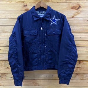 Könnte beinhalten: Marineblaue Jacke mit weißem Sternlogo auf der Brust. Die Jacke hat zwei Brusttaschen, einen Reißverschluss und geknöpfte Manschetten. Sie hängt vor einem Holzgrund.
