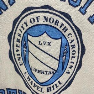 K&ouml;nnte beinhalten: Nahaufnahme eines wei&szlig;en Sweatshirts mit einem hellblauen und marineblauen Siegel der University of North Carolina Chapel Hill. Das Siegel enth&auml;lt die W&ouml;rter "University of North Carolina Chapel Hill", "LVX" und "Libertas".