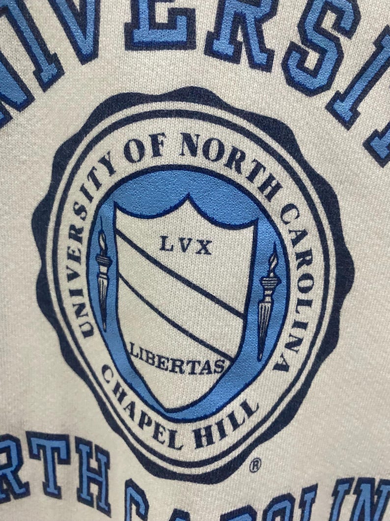 K&ouml;nnte beinhalten: Nahaufnahme eines wei&szlig;en Sweatshirts mit einem hellblauen und marineblauen Siegel der University of North Carolina Chapel Hill. Das Siegel enth&auml;lt die W&ouml;rter "University of North Carolina Chapel Hill", "LVX" und "Libertas".