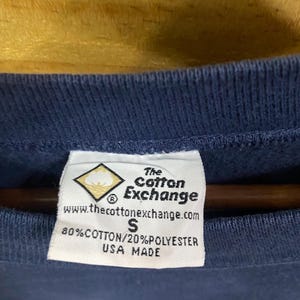 May include: Close-up of a navy blue garment tag. The tag is white with black text, including the brand "The Cotton Exchange" and the website address. It also states the material composition as 80% cotton and 20% polyester, and "USA MADE".