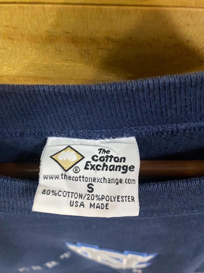 May include: Close-up of a navy blue garment tag. The tag is white with black text, including the brand "The Cotton Exchange" and the website address. It also states the material composition as 80% cotton and 20% polyester, and "USA MADE".