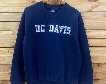May include: Navy blue crewneck sweatshirt with the text "UC DAVIS" in white block letters. The sweatshirt is hanging on a wooden hanger against a wood-paneled background. The sweatshirt has long sleeves and a ribbed collar and cuffs.
