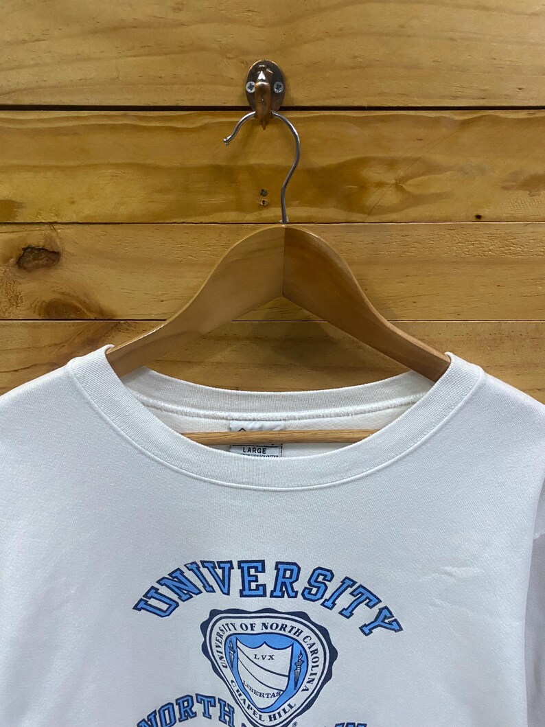 K&ouml;nnte beinhalten: Wei&szlig;es Sweatshirt mit Rundhalsausschnitt und dem blauen Schriftzug "UNIVERSITY OF NORTH CAROLINA". Das Sweatshirt hat eine Schildgrafik mit den W&ouml;rtern "CHAPEL HILL" und "LVX". Das Gr&ouml;&szlig;enetikett lautet "LARGE".