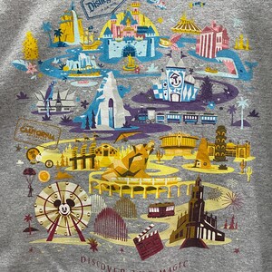 May include: A gray t-shirt with a colorful illustration of Disneyland park. The illustration includes iconic landmarks like Sleeping Beauty Castle, the Matterhorn, and the Haunted Mansion. The text "Discover the Magic" is printed at the bottom of the shirt.