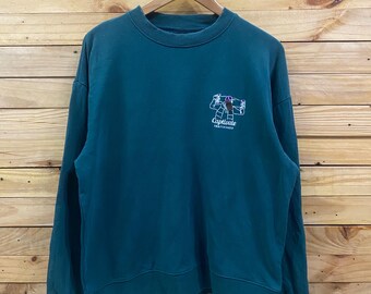 May include: A teal sweatshirt with a crew neck and long sleeves. The sweatshirt features a small graphic on the upper left chest with the text "Captivate Skateboards". The garment is hanging on a wooden hanger against a wooden background.
