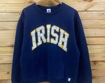 May include: Navy blue crewneck sweatshirt with the word "IRISH" arched across the chest in white and gold lettering. The sweatshirt is made by Russell Athletic. The background is a wooden wall.