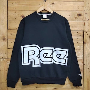 Op de afbeelding: Zwart sweatshirt met een wit Reebok-logo op de voorkant. Het logo is in een vet, blokkerig lettertype.