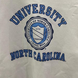 K&ouml;nnte beinhalten: Ein wei&szlig;es Sweatshirt mit den W&ouml;rtern "UNIVERSITY" und "NORTH CAROLINA" in blauen Blockbuchstaben. In der Mitte des Sweatshirts befindet sich ein kreisf&ouml;rmiges Emblem mit den Worten "UNIVERSITY OF NORTH CAROLINA CHAPEL HILL" um einen Schild.