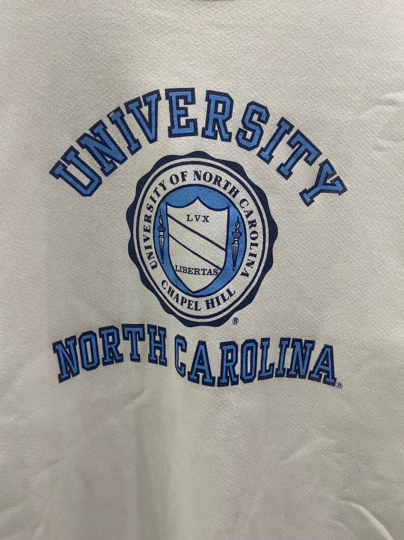 K&ouml;nnte beinhalten: Ein wei&szlig;es Sweatshirt mit den W&ouml;rtern "UNIVERSITY" und "NORTH CAROLINA" in blauen Blockbuchstaben. In der Mitte des Sweatshirts befindet sich ein kreisf&ouml;rmiges Emblem mit den Worten "UNIVERSITY OF NORTH CAROLINA CHAPEL HILL" um einen Schild.