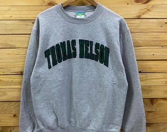 May include: A gray Champion sweatshirt with the text "THOMAS NELSON" in green block letters across the chest. The sweatshirt is hanging on a wooden hanger against a wooden background.