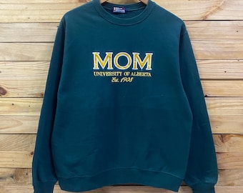 May include: A dark green crewneck sweatshirt featuring yellow embroidered text that reads MOM UNIVERSITY OF ALBERTA Est. 1908. The garment has long sleeves, a ribbed collar, and ribbed cuffs, hanging on a wooden hanger against a wood panel wall.