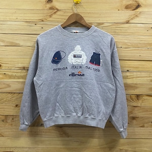 May include: Gray sweatshirt with a white embroidered logo that says "ellesse" and "ITALIA" with a white hooded jacket, a blue and white baseball cap, and blue and red shorts embroidered on the front. The text "PERUGIA DAL 1959" is also embroidered on the front.