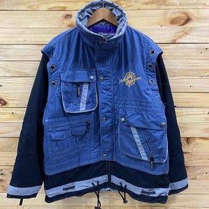 May include: A blue and black jacket with multiple pockets and a compass logo on the front. The jacket has a drawstring at the bottom and a zipper closure.