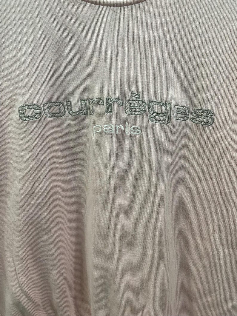 May include: Pale pink sweatshirt with the embroidered text "courr&egrave;ges paris" in a darker gray color. The fabric has a ribbed texture, and the stitching is visible.