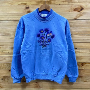 May include: A light blue sweatshirt with a high neck and long sleeves. The front features a floral design with blue flowers and two blue birds. The sweatshirt is hanging on a wooden hanger against a wooden background.