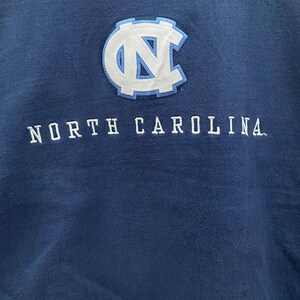 May include: Navy blue sweatshirt featuring the text "NORTH CAROLINA" in white block letters. Above the text is a white and light blue patch with the letters "NC" inside a circular design. The sweatshirt is made of a soft material.