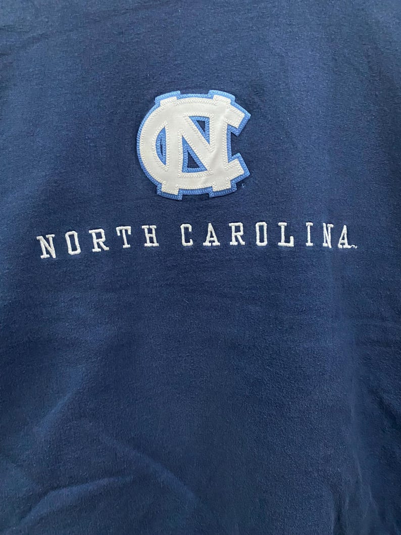 May include: Navy blue sweatshirt featuring the text "NORTH CAROLINA" in white block letters. Above the text is a white and light blue patch with the letters "NC" inside a circular design. The sweatshirt is made of a soft material.