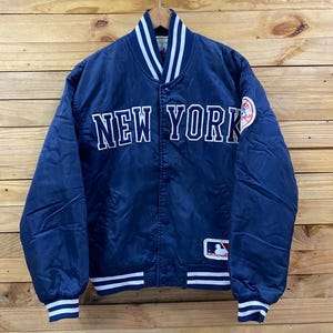 May include: A navy blue satin bomber jacket with the words "NEW YORK" in white block letters across the chest. The jacket has white and navy striped cuffs and collar, and a team logo patch on the sleeve. The jacket is hanging on a wooden background.