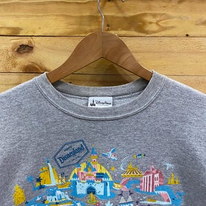 May include: Gray sweatshirt with a colorful graphic print of Disneyland park attractions. The text "Disneyland" is printed in a white box on the graphic.