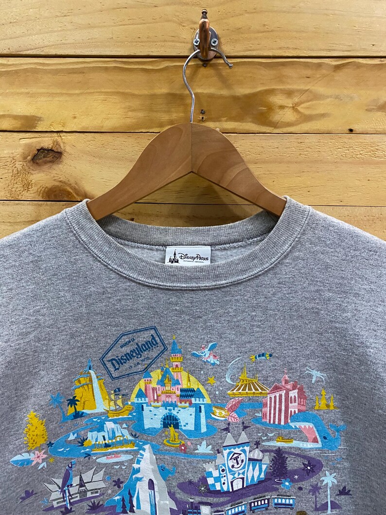 May include: Gray sweatshirt with a colorful graphic print of Disneyland park attractions. The text "Disneyland" is printed in a white box on the graphic.