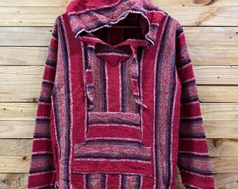 May include: A red and purple striped Baja hoodie with a hood and a front pocket. The hoodie is made of a woven fabric and has a drawstring at the neck. The background is a wooden wall.