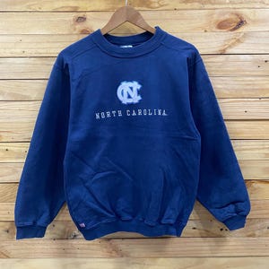 May include: Navy blue sweatshirt with the words "NORTH CAROLINA" in white lettering. The sweatshirt features a light blue and white logo on the chest. The sweatshirt is hanging on a wooden hanger.
